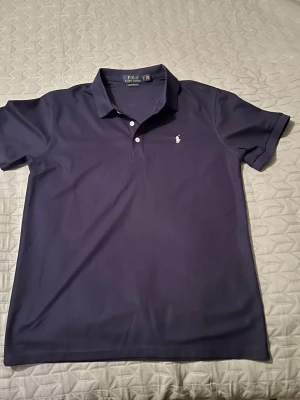 Mörkblå pikétröja från Polo Ralph Lauren - Snygg mörkblå pikétröja från Polo Ralph Lauren med klassisk krage och två knappar framtill. Tröjan har korta ärmar och den ikoniska broderade loggan på bröstet. Perfekt till jeans eller shorts för en clean look.