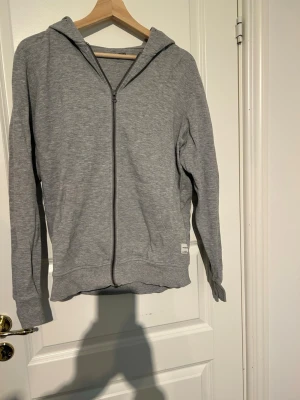 Grå hoodie från Jack & Jones - Säljer en stilren grå hoodie från Jack & Jones Essentials i storlek M. Hoodien har dragkedja framtill, huva och ribbade muddar vid ärmslut och nederkant. Tillverkad i mjukt bomullsmaterial som är skönt att ha på sig. Perfekt för en avslappnad look.