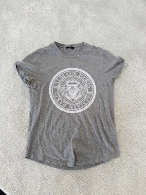 Grå t-shirt från Balmain Paris - Snygg grå t-shirt från Balmain Paris i storlek M, modellen är Slim fit. Stort vitt tryck framtill. Klassisk rund halsringning och t shirt armlängd. Inga slitningar, i fint skick.