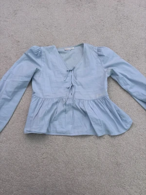 Ljusblå blus med volang och knyt - Supersöt ljusblå blus med v-ringning, knytband framtill och volang nedtill. Blusen har långa ärmar och lätt puff vid axlarna. Perfekt att styla med jeans eller kjol för en fräsch look.