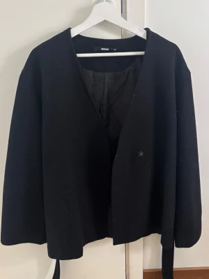 Svart kappa med bälte från Bikbok  - Stilren svart kappa från Bikbok med enkel knäppning och tillhörande bälte i midjan. Kappan har rak passform, V-ringad hals. Perfekt för dig som gillar minimalistisk och modern stil.
