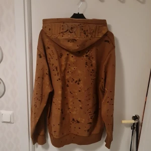 Brun hoodie från Supreme - Säljer en brun hoodie från Supreme med ett unikt stänkmönster. Hoodien har en stor ficka framtill och en broderad logga på huvan. Tillverkad i 100% bomull och gjord i Kanada. Perfekt för en avslappnad stil.
