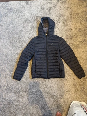 Svart pufferjacka från Hangten - Säljer en svart pufferjacka från Hangten med huva och dragkedja framtill. Jackan har quiltad design och är lätt vadderad, perfekt för kalla vinterdagar. Enkel och stilren look som passar till det mesta.