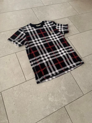 Svart rutig t-shirt med vita och röda detaljer - Säljer en svart t-shirt med stort rutigt mönster i vitt och rött. T-shirten har rund hals och korta ärmar. Perfekt för dig som vill sticka ut med en cool och trendig look. Materialet känns mjukt och bekvämt, troligtvis bomull eller polyester.