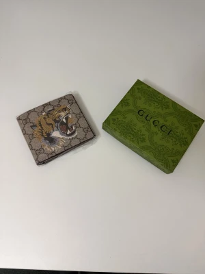 Plånbok med tigertryck - Snygg plånbok  i beige canvas med klassiskt GG-mönster och en stor broderad tiger på framsidan. Plånboken har en fyrkantig form och levereras med original grön. Perfekt accessoar för dig som vill sticka ut.