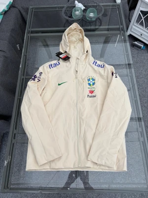 Ljusbeige lättviktsjacka / windbreaker med huva – L - Stilren ljusbeige/creamfärgad lättviktsjacka med huva. Tunt och följsamt material med luftig mesh på insidan. Hel dragkedja framtill och sportig, minimalistisk design. Perfekt som vindjacka, träningsjacka eller till vardags. Enkel att matcha med jeans, chinos, shorts eller träningskläder. Storlek L.