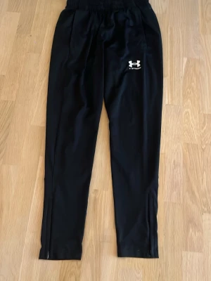 Svarta träningsbyxor Under Armour - Säljer ett par svarta träningsbyxor från Under Armour med logga på låret. Byxorna har resår i midjan och raka ben. Perfekta för gymmet eller löpning. Materialet är lätt och andas bra.