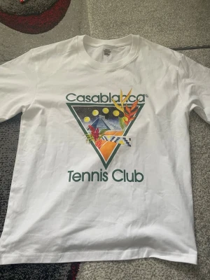 Vit Casablanca Tennis Club t-shirt - Säljer en vit t-shirt med färgglatt tryck från Casablanca Tennis Club. T-shirten har klassisk passform, korta ärmar och rund hals. Trycket har tropiska inslag och grafiska detaljer i grönt, gult och orange. Perfekt för dig som gillar sportig och trendig stil.