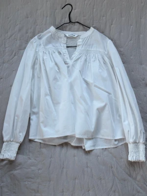 Vit blus från Soft Rebels XS - Stilren vit blus från Soft Rebels i storlek XS. Blusen har långärmade ärmar med smockade muddar, v-ringad hals med knappar och lätt rynkade detaljer upptill. Tillverkad i ett mjukt, vävt material som ger en luftig känsla.