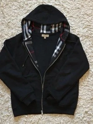 Svart hoodie med rutigt foder - Snygg slim fit svart zip up  och klassiskt rutigt foder i huvan. Hoodien har fickor framtill och justerbara snören i huvan. Perfekt för dig som gillar stilrena detaljer och vill ha något bekvämt och trendigt. Materialet känns mjukt och skönt mot huden.