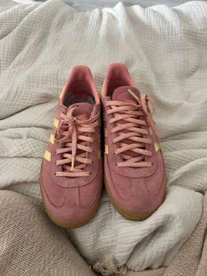 Adidas rosa Spezial sneakers - Säljer ett par Adidas Spezial sneakers i rosa mocka med ljusgula ränder och detaljer. Skorna har klassisk låg profil och gummisula som ger retrovibbar. Perfekta för dig som vill sticka ut med färg och stil. Utplpckad sula. Nypris 1200kr