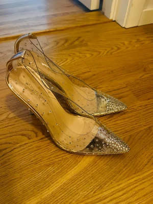 Transparenta pumps med strassdetaljer - Säljer ett par transparenta pumps från Marrio Rossi med spetsig tå och klack. Skorna är dekorerade med små glittrande strassdetaljer över hela ovandelen och har en elegant, modern look. Storlek 37 och endast använda en gång. 
