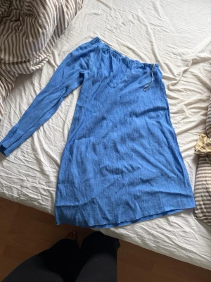 Blå one shoulder klänning - Snygg blå kortklänning med one shoulder-design och lång ärm. Klänningen har en avslappnad passform och är tillverkad i ett lätt material, perfekt för varma dagar. Enkel och stilren look som passar till många tillfällen.