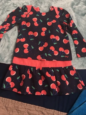 Mörkblå körsbärsklänning Little Marc Jacobs - Supersöt mörkblå klänning från Little Marc Jacobs med färgglatt körsbärsmönster och kontrasterande korallröda detaljer. Klänningen har lång ärm, rund hals och en volangkjol. Perfekt för dig som gillar lekfulla prints och färg!