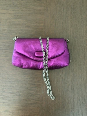 Lila metallic kuvertväska med kedja - Snygg kuvertväska i metallic-lila med silverfärgad kedjerem. Väskan har en mjuk, glansig yta och ett lock med enkel stängning. Perfekt accessoar för att lyfta din outfit med en färgklick och trendig känsla.