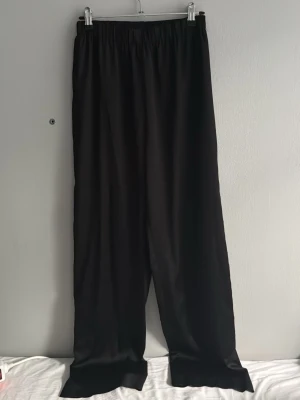 Svarta satinbyxor med resårmidja - Säljer ett par svarta leggings med breda, raka ben och bekväm resår i midjan. Materialet ser ut att vara mjukt och stretchigt, perfekt för en avslappnad och trendig look. Passar dig som gillar minimalistisk stil.