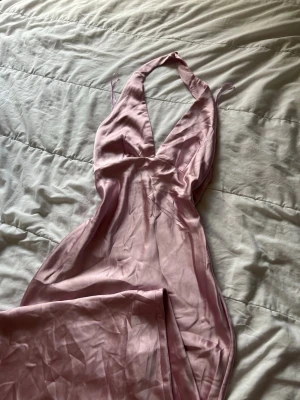Ljusrosa långklänning i satin - Säljer en elegant ljusrosa långklänning i satin med djup v-ringning och halterneck. Klänningen har en slits framtill och ett mjukt, glansigt material som ger en lyxig känsla. Perfekt för speciella tillfällen och sommarens fester.