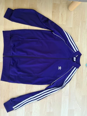 Lila Adidas track jacket med vita ränder - Säljer en klassisk lila Adidas track jacket med dragkedja framtill och vita ränder längs ärmarna. Jackan har ribbade muddar vid ärmslut och nederkant, två sidofickor och Adidas logga broderad på bröstet. Materialet är troligtvis polyester, perfekt för sport eller streetwear.