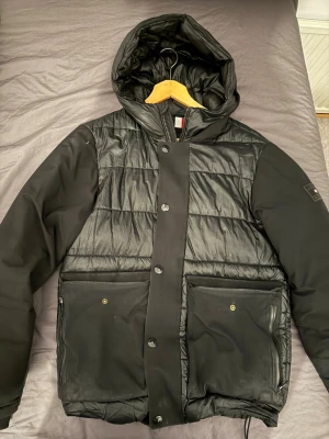 Vinterjacka Tommy Hilfigher - Svart jacka med huva och dragkedja framtill. Jackan har två fickor med lock och knappar samt två sidofickor med dragkedja. Ärmarna har en slät design medan bålen är vadderad med horisontella sömmar.   Obs: finns en liten öppning i vänster ärm, se sista bilden.