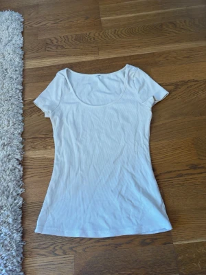 Vit ribbad kortärmad topp - Säljer en enkel och fräsch vit topp med ribbad struktur och korta ärmar. Toppen har en rund, lite djupare urringning och är figurnära i modellen. Perfekt att styla med jeans eller kjol för en clean look.