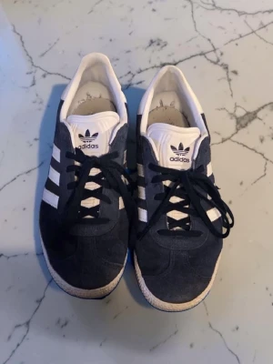 Adidas Gazelle mörkblå sneakers - Mörkblå Adidas Gazelle sneakers med klassiska vita ränder på sidorna och vit plös med Adidas-logga. Ovandelen är i mocka och sulan är vit med gummidetaljer. Skorna har svarta skosnören och låg profil.
