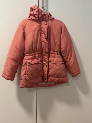 Jacka  - Snygg rosa pufferjacka från KappAhl med huva och dragkedja samt tryckknappar framtill. Jackan har två stora fickor med lock och guldiga knappar, samt resår i midjan för en skön passform. Fodrad med mjukt tyg för extra värme.