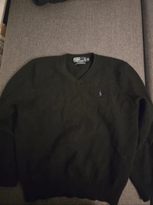 Svart stickad tröja från Polo Ralph Lauren - Klassisk svart stickad tröja från Polo Ralph Lauren med lila broderad logga på bröstet. Tröjan har V-ringning, långa ärmar och ribbade muddar. Perfekt för dig som gillar stilrena och tidlösa plagg i garderoben. Size XL but fits S-M 100% wool