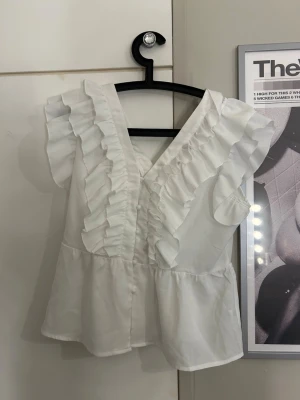 Vit blus med volanger och v-ringning - Snygg vit blus med v-ringning och volangdetaljer längs bröstet och axlarna. Blusen har korta ärmar och en lätt utsvängd passform. Perfekt för dig som gillar en romantisk och trendig stil. Materialet känns lätt och luftigt. Perfekt festtopp 🦄🦄