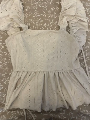 Vit blus med volang och spetsdetaljer - Söt vit blus med volangkant och broderade spetsdetaljer. Blusen har breda axelband med volanger och knytband i sidan. Perfekt för dig som gillar romantisk och feminin stil.