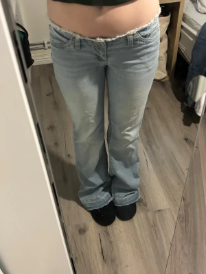Ljusblå bootcut jeans med rå kant - Säljer ett par ljusblå bootcut jeans med låg midja och rå, fransad kant upptill. Jeansen har klassisk femficksdesign och är tillverkade i denim med lite stretch. Perfekta för en avslappnad och trendig look. Säljer då de är för stora, lite upptrampade i bak