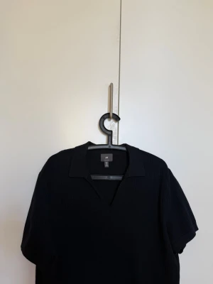 Svart ribbad polo från H&M - Säljer en svart ribbad polotopp från H&M med korta ärmar och krage. Toppen har en v-ringad öppning framtill och är gjord i ett mjukt, stretchigt material som sitter snyggt på kroppen.