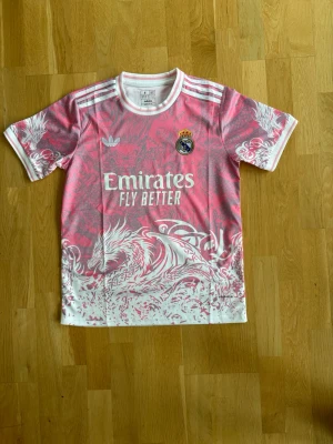 Rosa Real Madrid tröja Adidas - Säljer en rosa Real Madrid tröja från Adidas med vitt drakmönster och vita detaljer. Tröjan har klubbmärke, Adidas-logga och 'Emirates Fly Better' tryckt på bröstet. Rund halsringning och korta ärmar. Perfekt för dig som älskar fotboll och unik design.