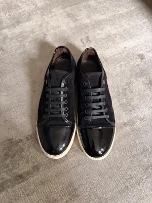 Svarta sneakers med lackdetaljer - Snygga svarta sneakers med coola lackade tåpartier och vita sulor. Skorna har svarta skosnören och ovandel i mocka eller textil, vilket ger en schysst kontrast till den blanka tån. Perfekta för dig som vill ha en stilren men ändå unik look. Storlek 46!