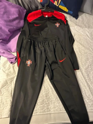 Svart och röd Nike träningsdress - Säljer en svart träningsdress från Nike med röda detaljer och klubbmärke på både jacka och byxor. Dressen har dragkedja vid benslut, elastisk midja och halv dragkedja på tröjan. Perfekt för träning eller uppvärmning. Materialet är troligtvis polyester.