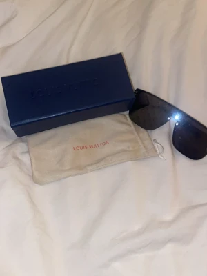 L V solglasögon - Snygga svarta solglasögon med spegelglas och ikoniskt mönster över hela linsen. Modellen har en futuristisk, bred sköldform och kommer med originalfodral och box. Perfekt accessoar för dig som vill sticka ut.