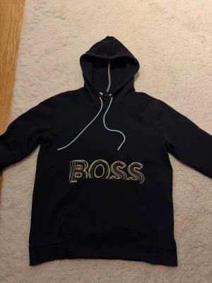 Svart hoodie med BOSS-tryck - Snygg svart hoodie med stort BOSS-tryck i vitt och gult på bröstet. Klassisk modell med huva och dragsnören samt en stor magficka framtill. Perfekt för en avslappnad och trendig stil.