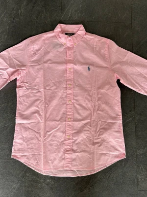 Rosa skjorta från Polo Ralph Lauren - Klassisk rosa skjorta från Polo Ralph Lauren med button-down krage och broderad logga på bröstet. Skjortan har långa ärmar, knappar framtill och är tillverkad i bomull. Perfekt för en clean och stilren look.