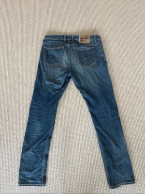 Jacob Cohën jeans - Ett par blå jeans med tvättad look och slitningar, inklusive hål på båda knäna. Klassisk femficksmodell med raka ben och knappgylf. Jeansen har en avslappnad vibe och passar perfekt till en streetwear-stil.