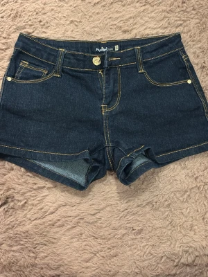 Mörkblå jeansshorts med guld detalj - Snygga mörkblå jeansshorts med klassiska fem fickor och kontrasterande gula sömmar. Modellen har normal passform och stängs med knapp och dragkedja. Perfekta för varma dagar och enkla att matcha med allt. Säljer pga den inte används