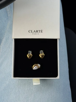 Guldiga hjärtörhängen & ring med stenar - Säljer detta superfina smyckes sett från Clarté Paris! Vattentåliga och ”Hypoallergenic”. Ringen är 16,5 mm (size 6). Använda en gång och säljer pga att jag är en silver tjej☀️  Köpt för 594kr❤️