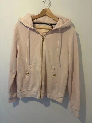 Ljusrosa hoodie med dragkedja - Säljer en ljusrosa hoodie med dragkedja framtill och två fickor med knappdetaljer. Hoodien har huva med dragsnören. 
