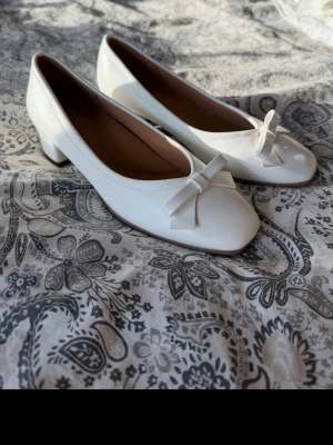 Vita ballerinaskor med rosett - Säljer ett par vita ballerinaskor med låg klack och söt rosett framtill. Skorna har rundad tå och är tillverkade i ett glansigt material som ger en stilren look. Perfekta för dig som gillar klassisk och feminin stil.
