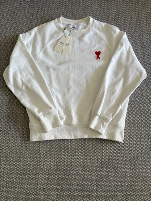 Vit Ami Paris tröja med rött hjärta - Vit Ami Paris tröja med rund hals och ribbade muddar. På bröstet finns en broderad röd detalj i form av ett hjärta och bokstaven A. Perfekt för en avslappnad och stilren look.