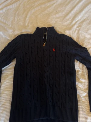 Ralph Lauren tröja - Mörkblå kabelstickad tröja från Polo Ralph Lauren med halv dragkedja och hög krage. Tröjan har ett rött broderat logotyp på bröstet och ribbade muddar. Perfekt för kalla dagar och snygg till jeans eller chinos.
