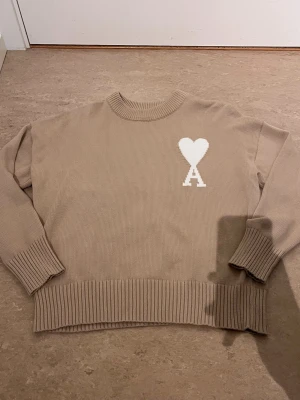Beige stickad tröja från Ami - Säljer en beige stickad tröja från Ami Alexandre Mattiussi med rund halsringning och ribbade muddar. Tröjan har ett vitt hjärta med bokstaven A broderat på bröstet. Perfekt för dig som gillar stilrena och trendiga plagg.