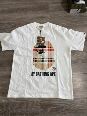Vit By Bathing Ape t-shirt - Snygg vit t-shirt från By Bathing Ape med stort tryck framtill i beige, svart och rött. Klassisk rund hals och korta ärmar. T-shirten har ett rutigt mönster i trycket och är tillverkad i mjuk bomull. Perfekt för en trendig streetwear-look.