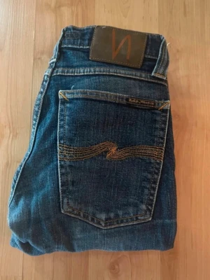 Nudie jeans  - Mörkblå jeans från Nudie Jeans, tajt long John. Midja 32cm, längd 99cm. OBS jeansen har hål på ena låret och vid knäna - se bild. JAG SKICKAR EJ FLER BILDER NÄR JAG BÄR JEANSEN! för bättre inblick i passformen rekommenderar jag att googla på modellnamnet. För storleksguide kolla måtten noggrant då jeansen alltid kan vara uppsydda heller krympta och jag ej tar emot returer :) s6,3