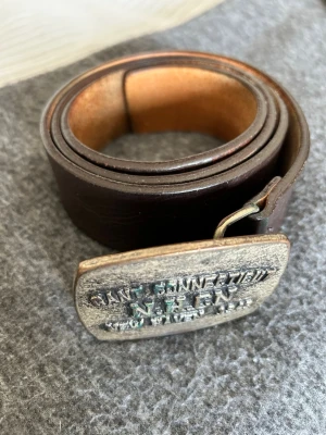 Brunt skärp med metallspänne från Gant - Snyggt brunt skärp från Gant med ett robust metallspänne där det står 'Gant Connecticut N.H.CN New Haven 1949'. Skärpet är gjort i skinn och har en klassisk, tidlös look som passar till jeans eller chinos.