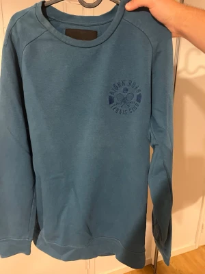 Blå sweatshirt från Björn Borg - Snygg blå sweatshirt från Björn Borg med rund halsringning och tryck på bröstet med texten 'Björn Borg Tennis Club' och två tennisrack. Perfekt för en sportig och avslappnad stil. Pris kan diskuteras vid snabb affär