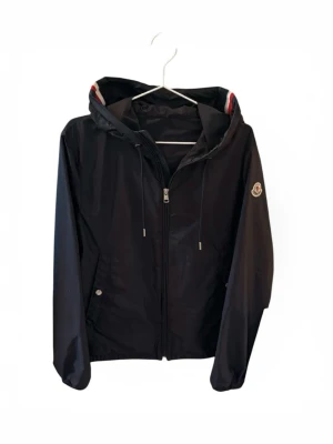 moncler windbreaker  - Snygg svart vindjacka från Moncler med dragkedja framtill, huva med röd och vit rand samt logotyp på ärmen. Jackan har dragsko i huvan och fickor med knappstängning. Perfekt för blåsiga dagar och enkel att matcha till olika outfits.